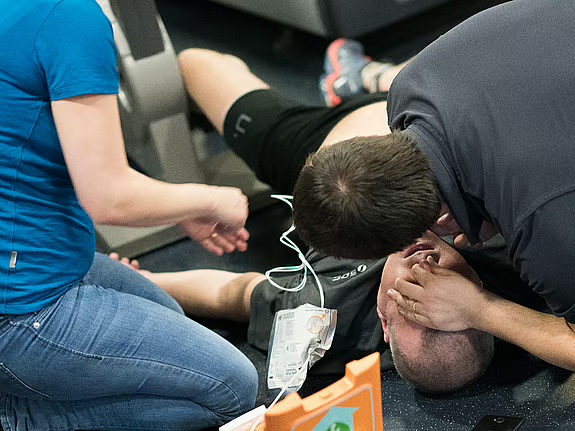 Två personer hjälper en man med hjärtstopp på ett gym, hjärtstartaren kopplas