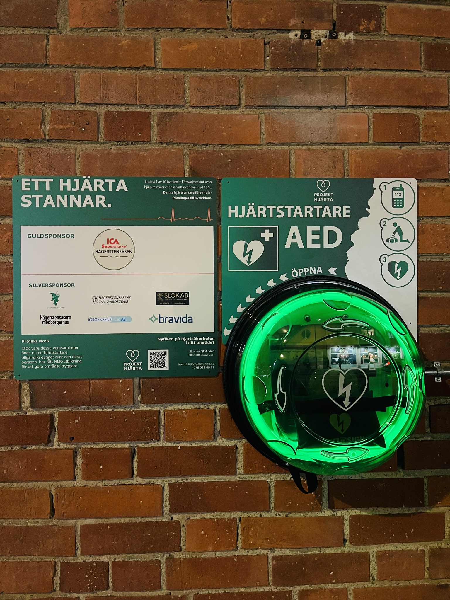Hjärtstartarkiosk och sponsortavla på en tegelfasad, upplysta i mörkret