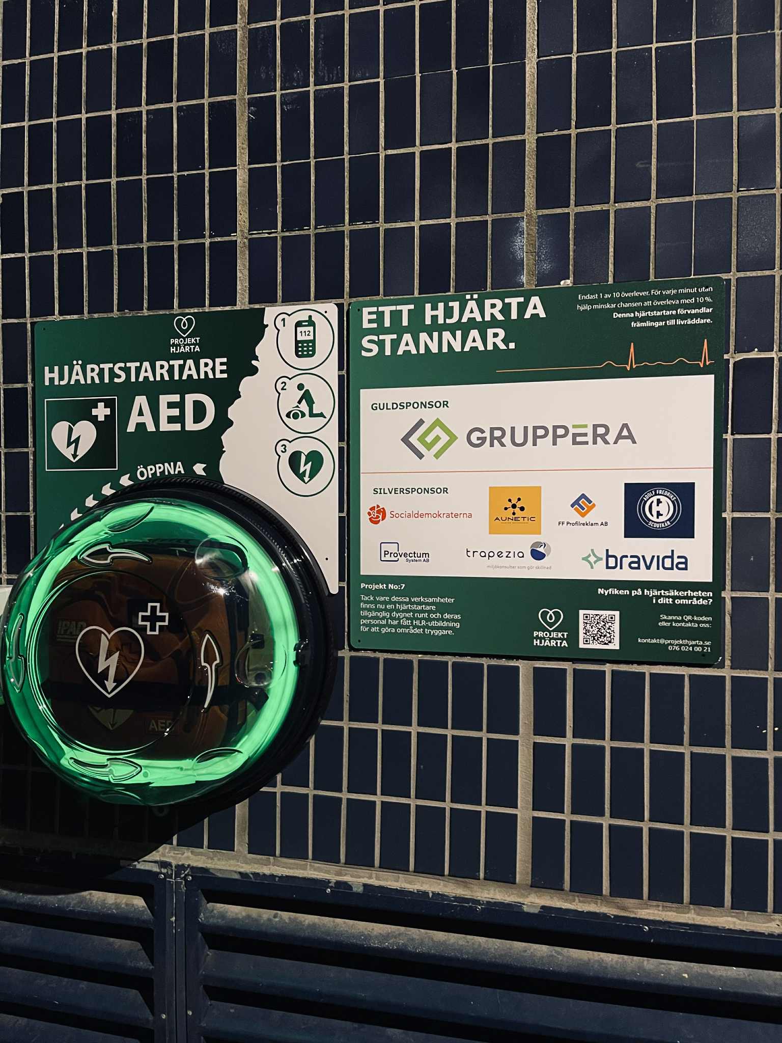 Hjärtstartarkiosken lyser grönt bredvid sponsortavlan på kvällen