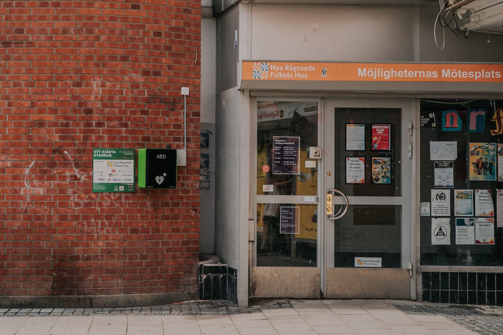Grön hjärtstartare i utomhusskåp på en tegelvägg, bredvid sponsorskylten vid Nya Rågsveds Folkets Hus