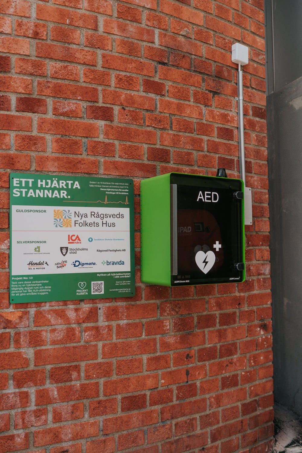 Sponsorskylt med företagsloggor bredvid en grön hjärtstartare på en tegelvägg